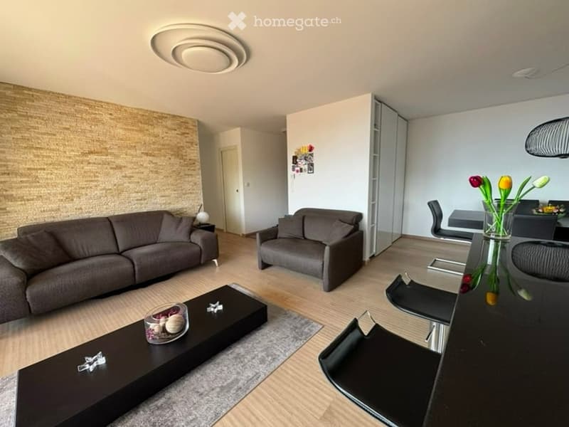 Modern living and dining area with a grey sofa, armchair, coffee table, dining table, chairs, and a vase with flowers on the counter.