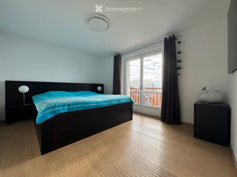 Modern bedroom with wooden flooring, large window with dark curtains, bed with blue bedding, nightstands with lamps, and a decorative mirror on a side table.