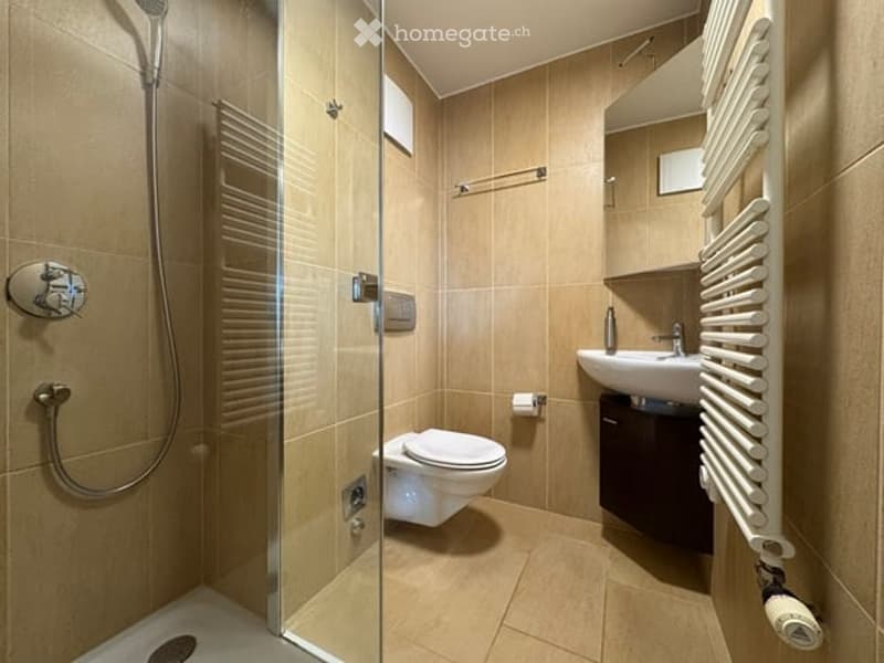 Modern bathroom with beige tiles, glass-enclosed shower, toilet, sink, and a white radiator.