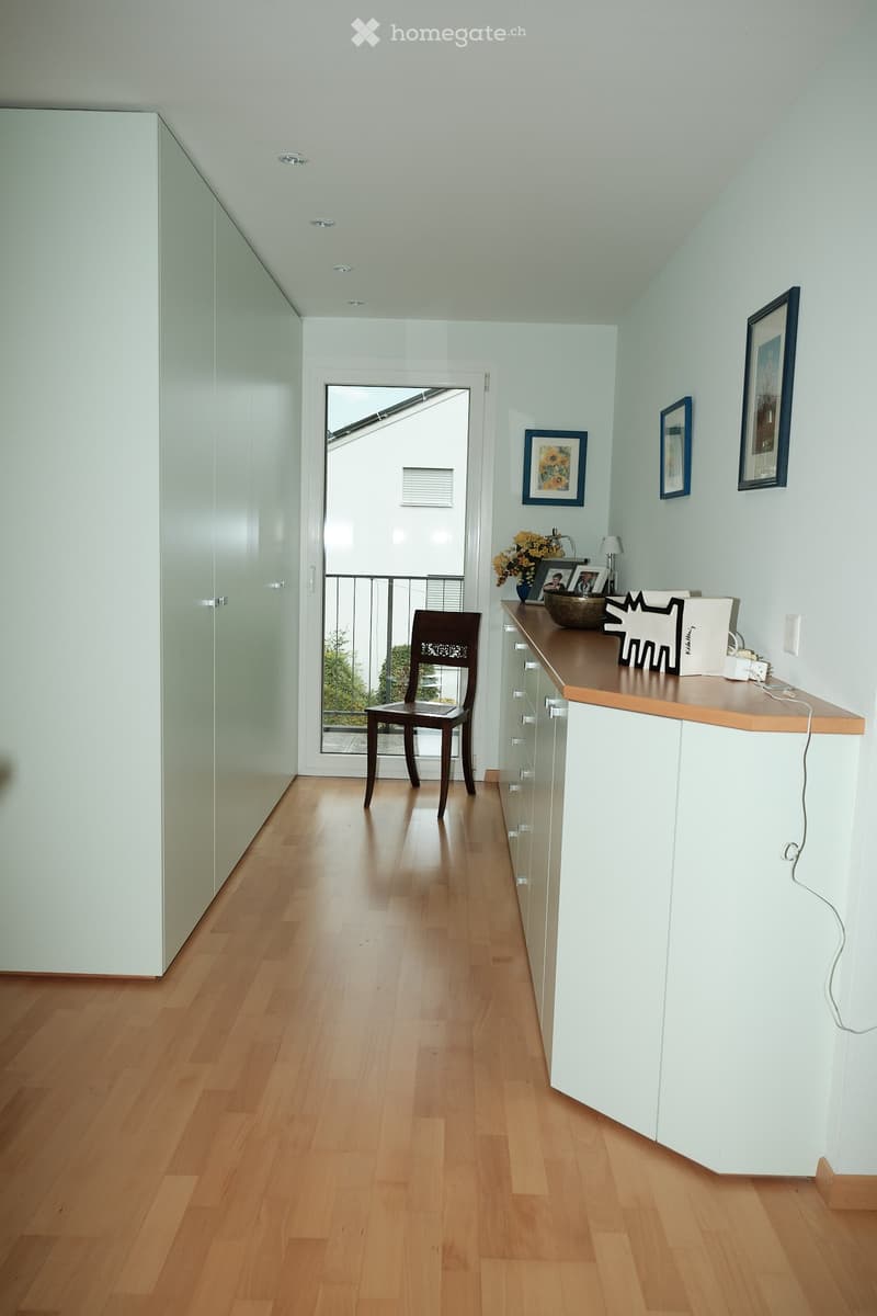 A modern hallway with light wooden flooring, white walls, a small table with a vase of flowers, a chair, and framed pictures on the wall. A door leads to a balcony.