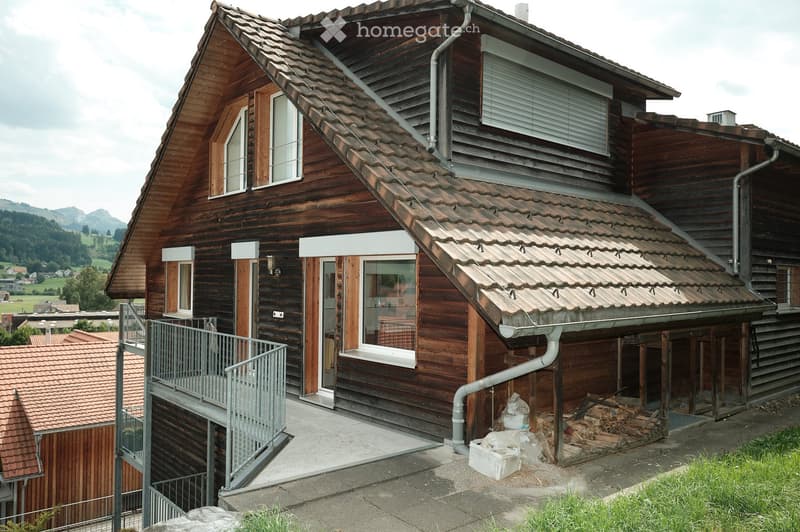 Two-story wooden house with a steeply pitched roof, multiple windows, and a balcony on the upper level. The house features a mix of wooden and concrete elements, with a visible chimney and a small stack of firewood near the entrance.
