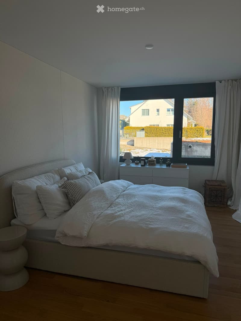 Modern bedroom with a large bed, white bedding, nightstand with lamp, and large windows with curtains overlooking a residential area.