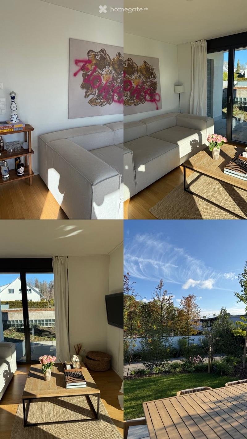 Modern living room with a grey sectional sofa, coffee table, side table, floor lamp, and artwork. Large windows offer a view of a well-maintained garden with a wooden outdoor table and chairs.