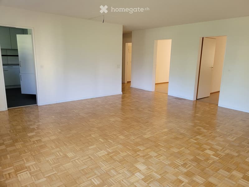 Empty room with light-colored walls and a herringbone-patterned wooden floor. Three doors lead to adjacent spaces, and a small kitchen area is visible through one doorway.
