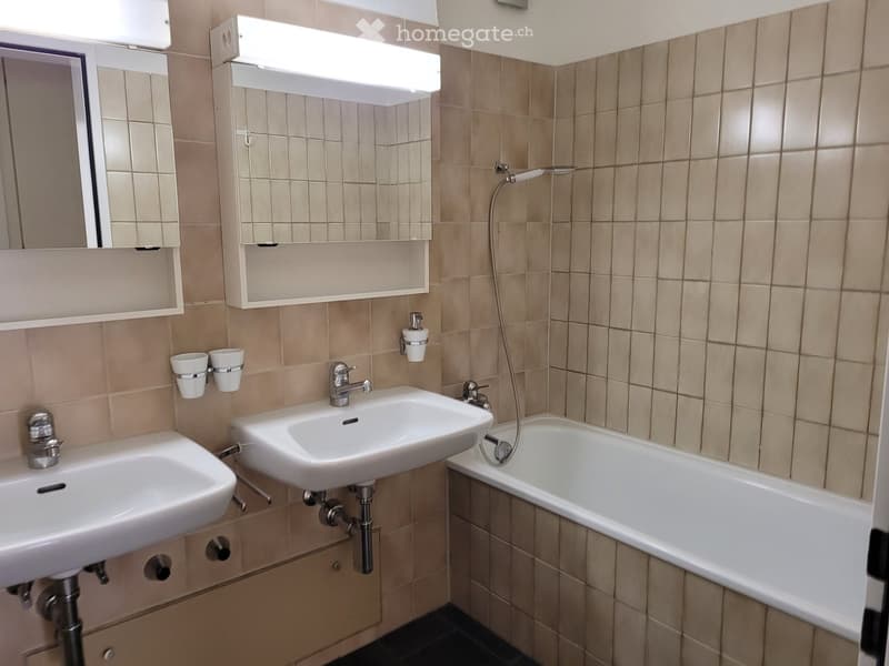 Bathroom with two sinks, a bathtub, a mirror, and a shower hose. Beige tiled walls and floor.