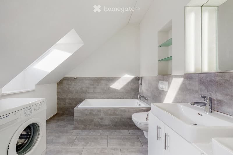 Modern bathroom with a bathtub, sink, toilet, washing machine, and a large mirror with built-in storage. The space features grey tiled walls and a skylight window providing natural light.