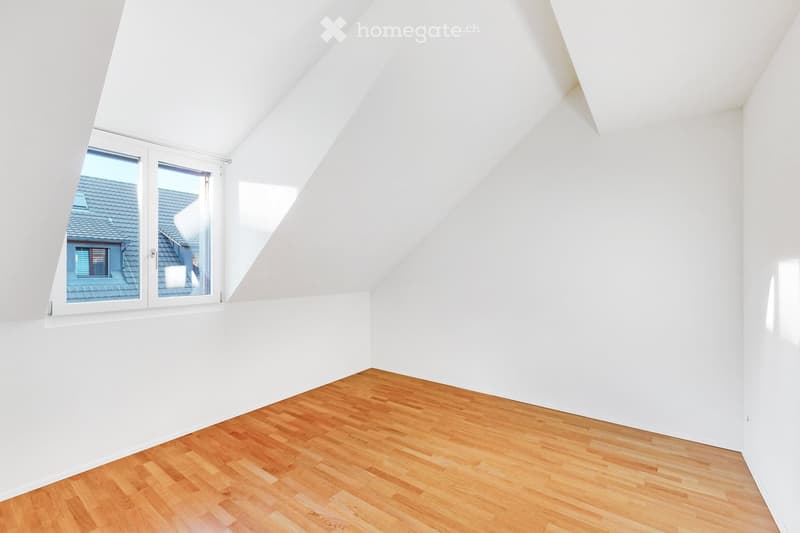 An empty attic room with white walls, wooden flooring, and a large skylight window offering a view of the roof and surrounding area.