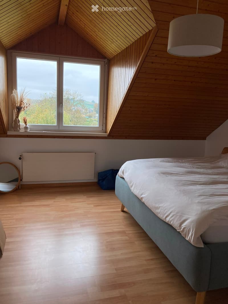 Bright attic bedroom with wooden floors, slanted wooden ceiling, large window offering a view, a bed with white linens, a vase with dried plants, a round mirror, and a radiator.