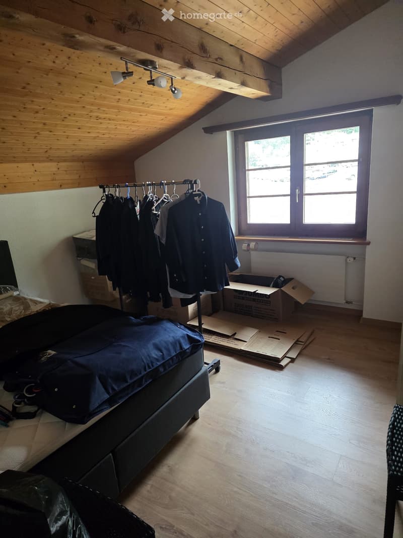 Bedroom with wooden floor, slanted wooden ceiling, large window, bed with dark blue cover, clothing rack with jackets, and cardboard boxes on floor.