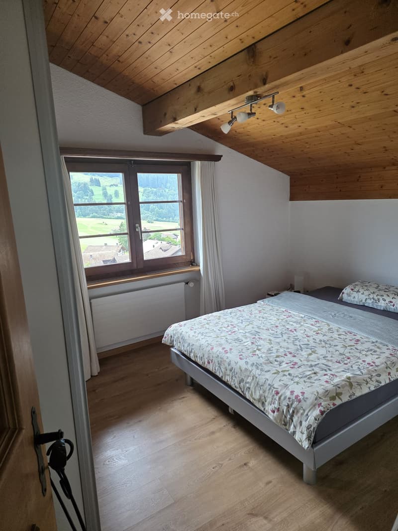 Cozy attic bedroom with wooden slanted ceiling, large window offering scenic view, single bed with floral bedding, radiator, and white curtains.