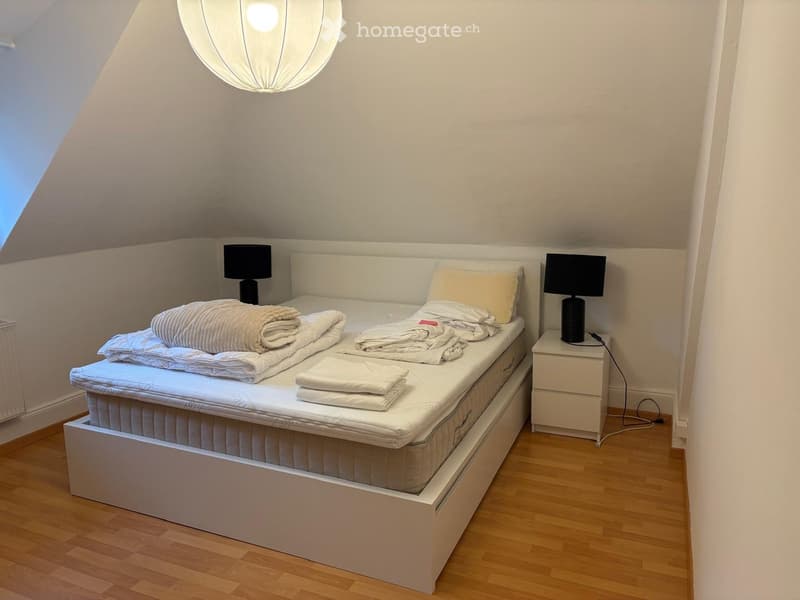 Cozy bedroom with a single bed, neatly made with folded blankets and pillows, a nightstand with a lamp, and warm lighting from a hanging spherical lamp.