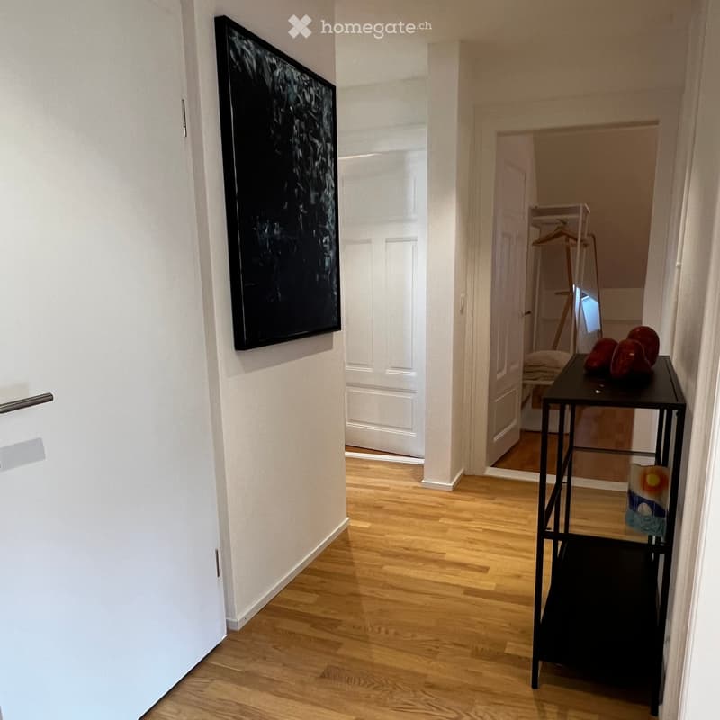 A well-lit hallway with wooden flooring, white walls, and a black artwork on the left wall. A small black table with decorative items is visible on the right side.