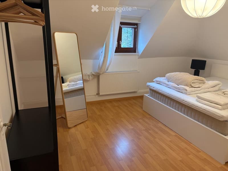 Cozy attic bedroom with wooden flooring, a neatly made bed, a full-length mirror, and a window with white curtains.