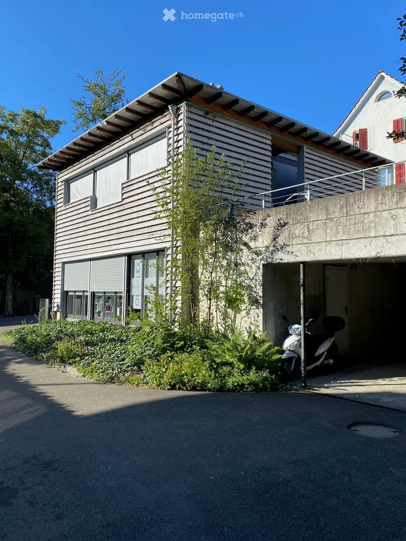 Modern two-story building with a flat roof, large windows, and a small balcony. A scooter is parked in the garage entrance.
