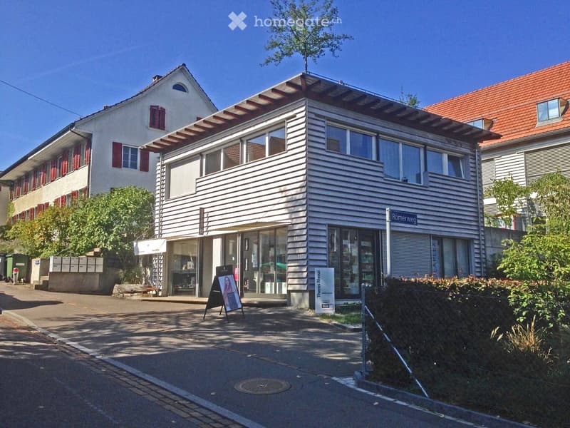 Two-story building with a modern design, featuring large windows, a flat roof, and a storefront with glass doors. The building has a signboard and is situated on a street with a sidewalk and a street sign.