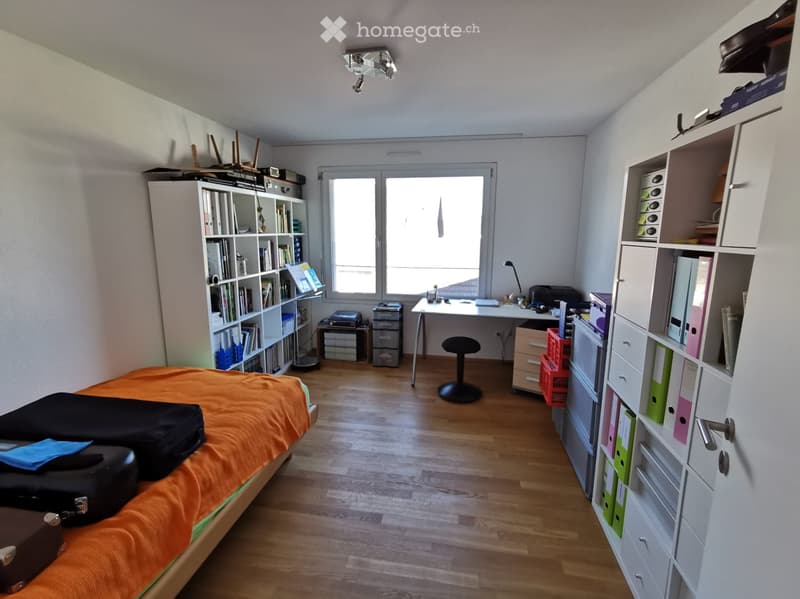 A well-lit bedroom with a wooden floor, a bed with an orange blanket, a desk with a chair, a bookshelf, and storage boxes.