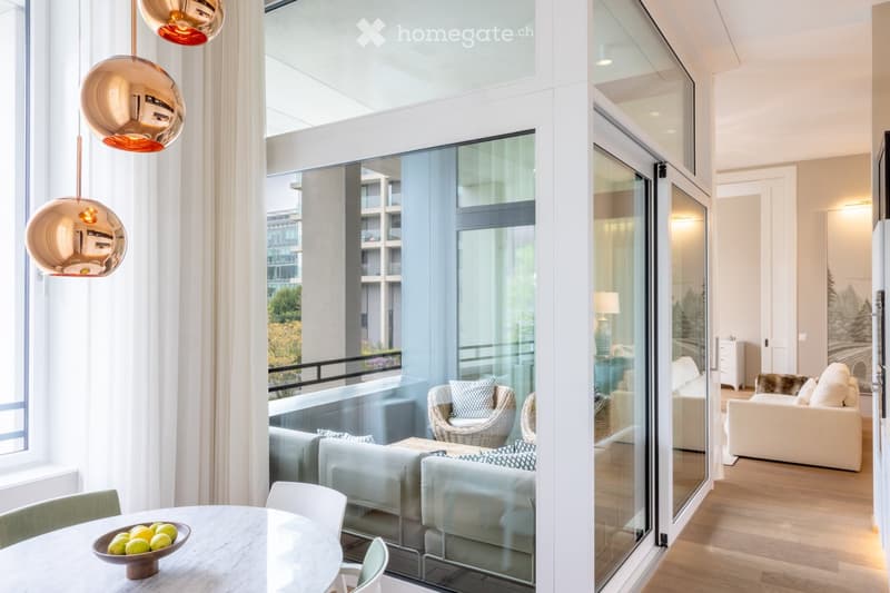 Modern living and dining area with large windows and a balcony. Features a round glass dining table with chairs, a white sofa, and pendant lights.