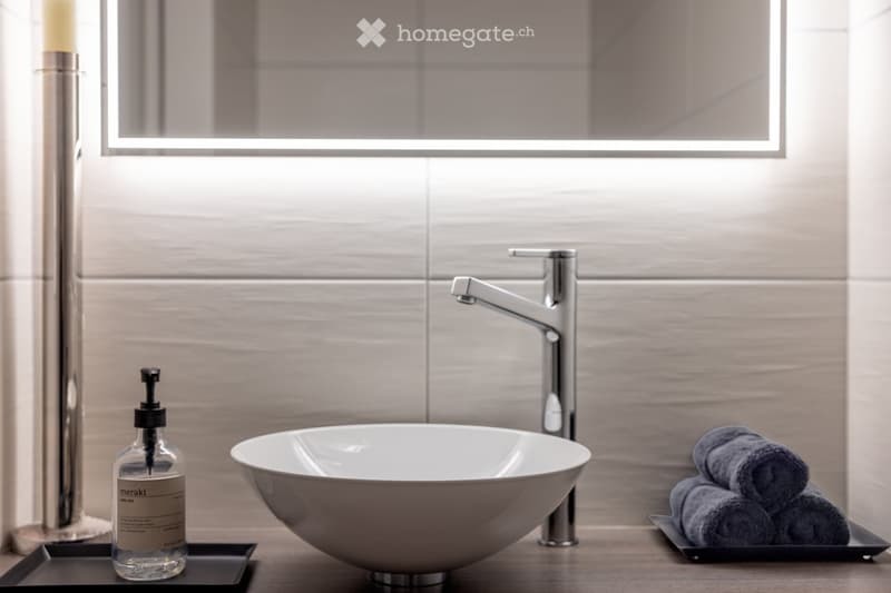 Modern bathroom with a sleek white sink, chrome faucet, soap dispenser, and neatly folded towels on a tray.