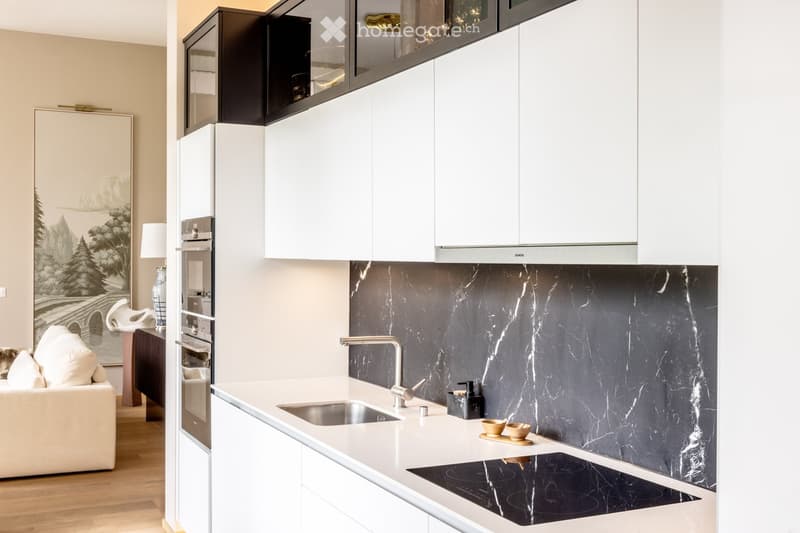 Modern kitchen with white cabinets, marble backsplash, stainless steel appliances, and a small living area with a sofa and table.