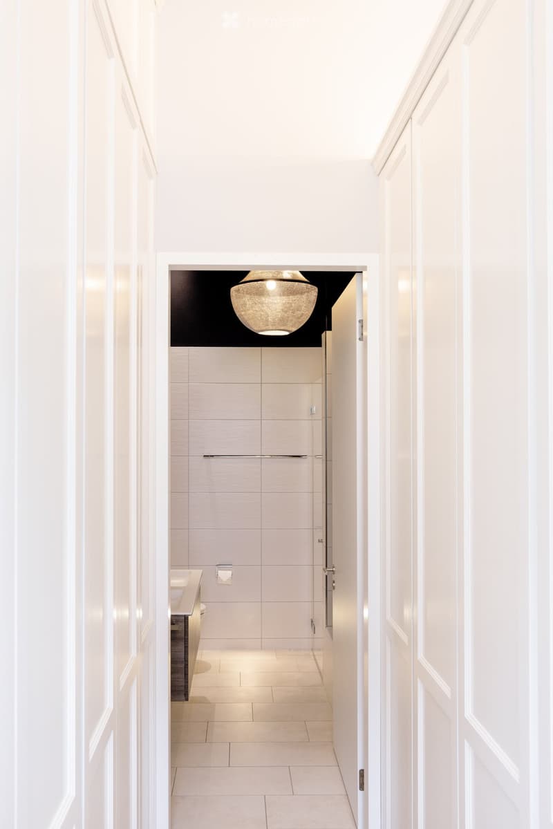 A modern, well-lit hallway with white walls and a sleek design, leading to a bathroom with a sink, toilet, and modern fixtures.