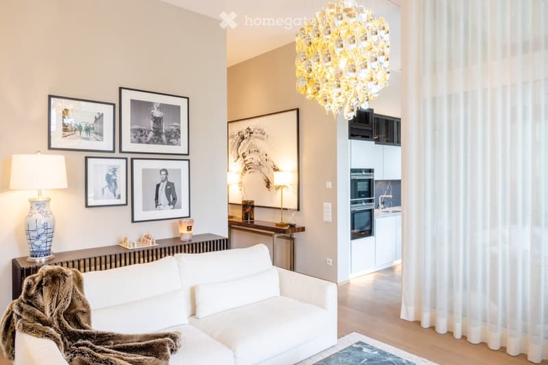 Modern living room with white sofa, fur throw, decorative table, framed pictures, and a chandelier. Adjacent kitchen area with modern appliances.