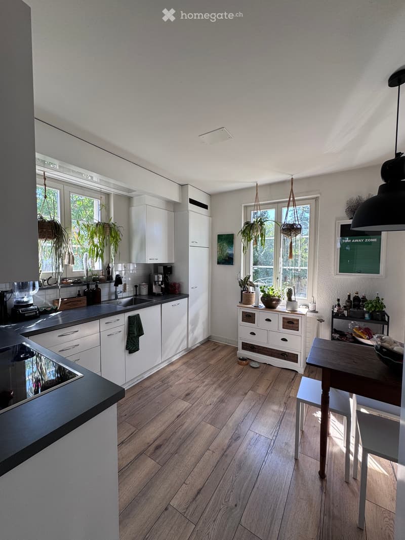 Modern kitchen with white cabinets, wooden flooring, and large windows. Features a sink, coffee maker, blender, oven, and a dining area with a table and chairs.