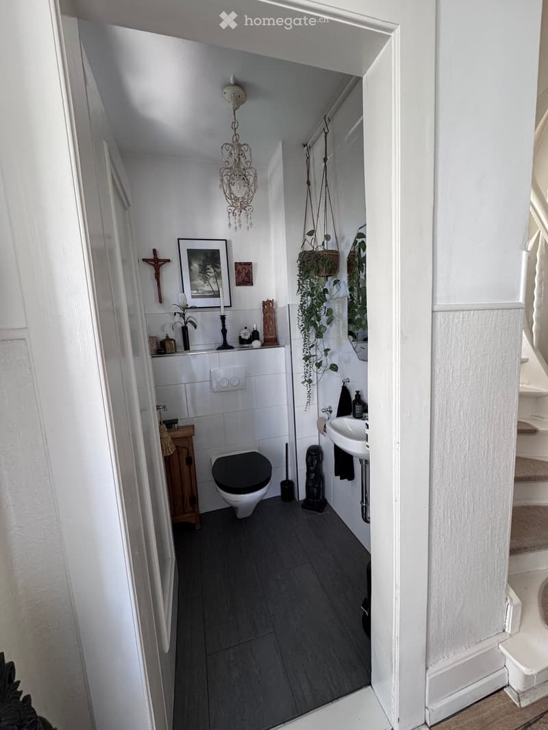 Modern bathroom with dark grey tiles, white walls, a toilet, sink, mirror, framed picture, candle, plant, wooden cabinet, and a chandelier.