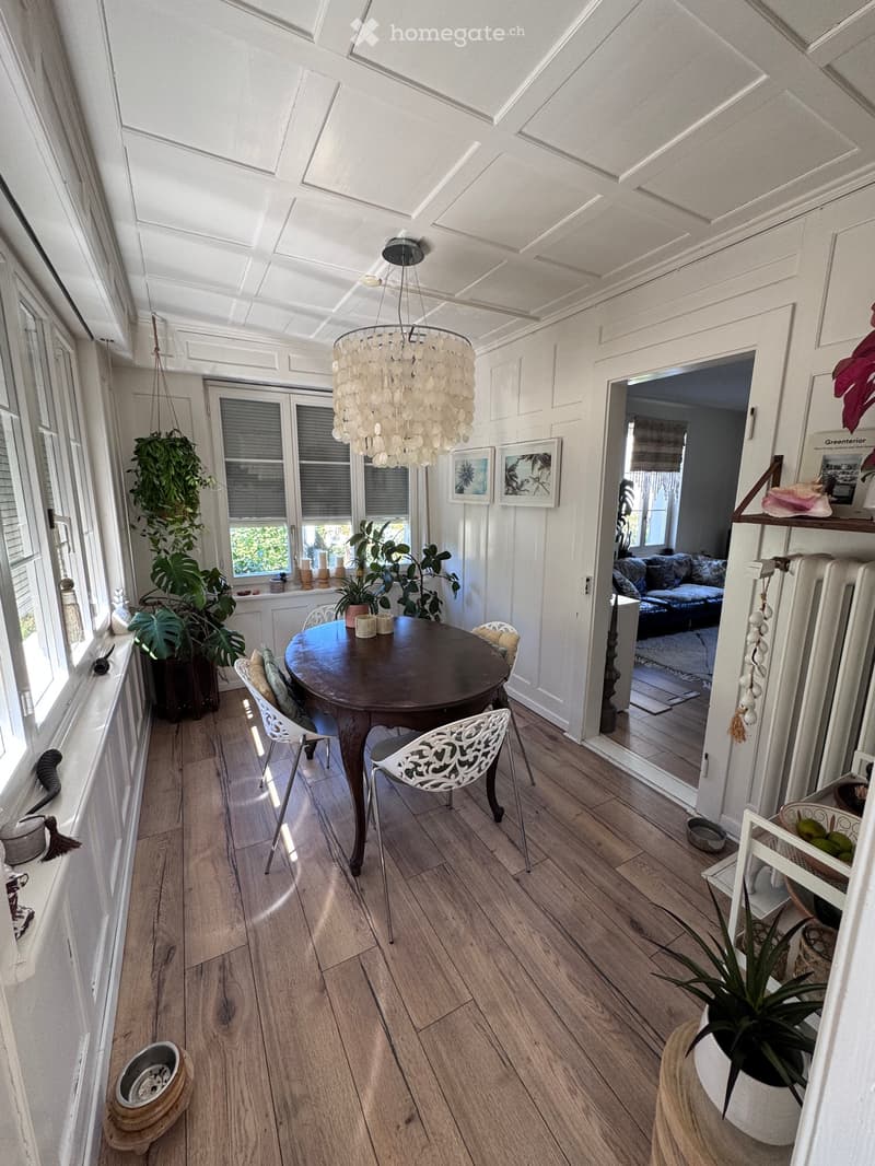Brightly lit room with wooden flooring, a round wooden table with four white chairs, a large chandelier, several potted plants, and picture frames on the wall. A doorway leads to another room with a sofa visible.