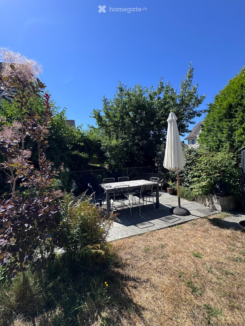 Garden with a patio area featuring a table, chairs, and an umbrella, surrounded by greenery and trees.