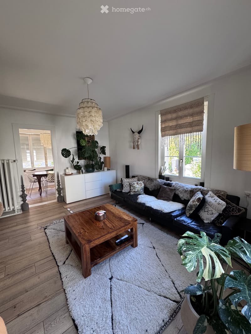 Modern living room with a black leather sofa, wooden coffee table, side table, and large windows with blinds. Features a white shag rug, pendant light, and decorative plants.
