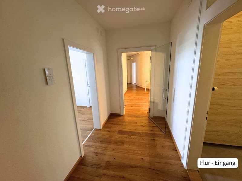 A clean, well-lit hallway with wooden flooring and white walls, featuring several open doors leading to other rooms.