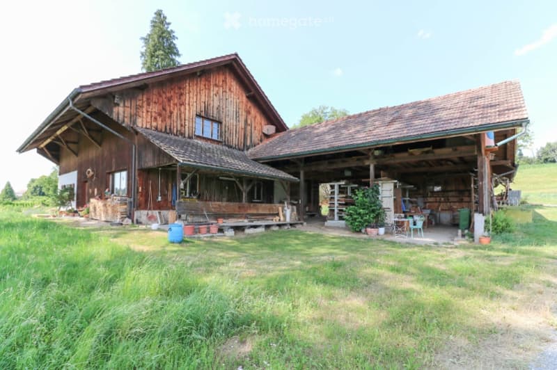 A rustic wooden farmhouse with a large barn attached, surrounded by a grassy yard with scattered outdoor furniture and potted plants.