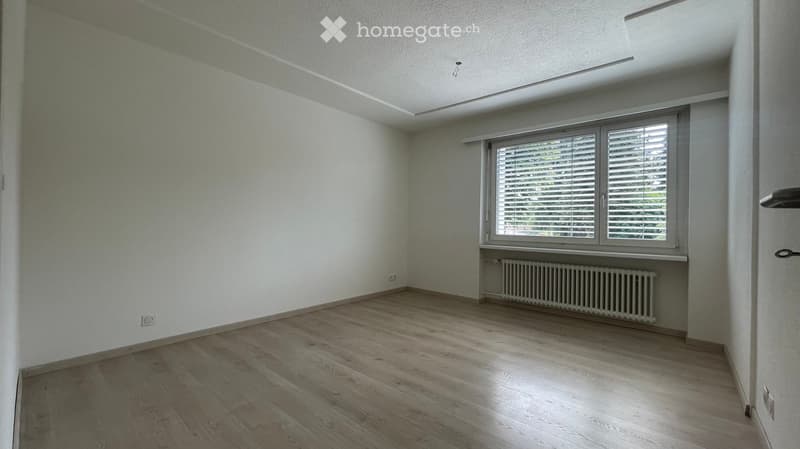 Empty room with light wooden flooring, white walls, and a large window with blinds. A radiator is visible below the window.