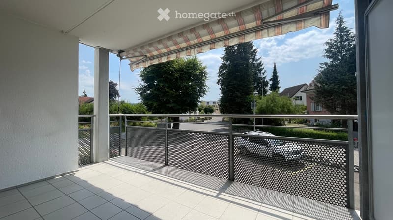 Balcony with tiled floor, metal railing, striped awning, and view of a residential area with trees and houses.