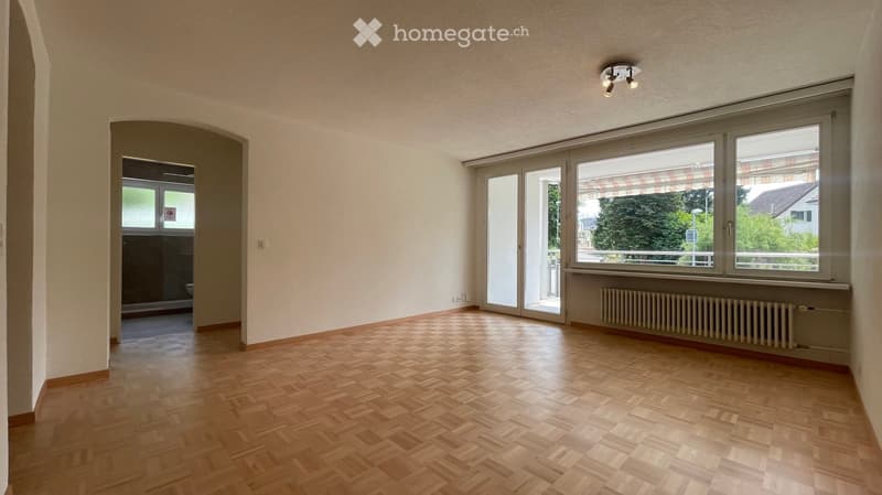 Empty room with wooden herringbone flooring, large windows, sliding glass doors, and a radiator. No furniture or equipment present.