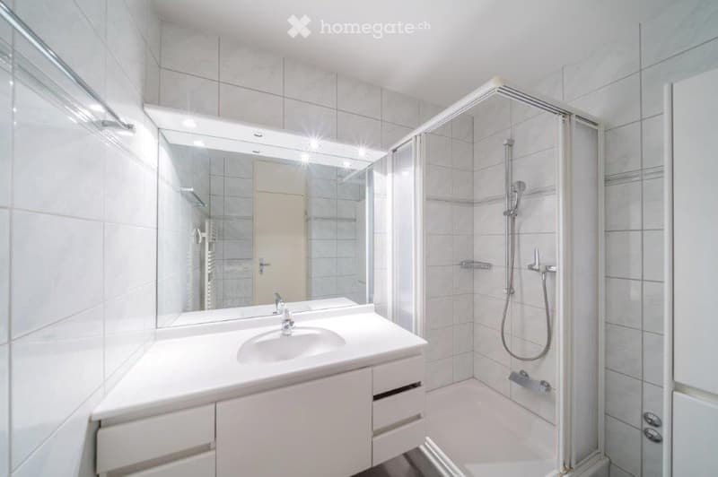 fully tiled white bathroom, walk-in shower with large mirror, white vanity with under cabinet drawers, towel rack, and a washing machine