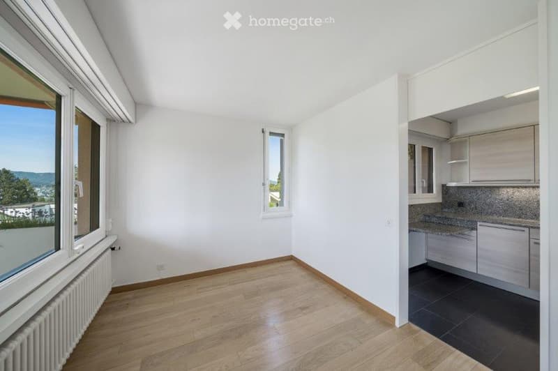 an empty room with large windows, hardwood floors, a small kitchenette, a radiator, and a small balcony