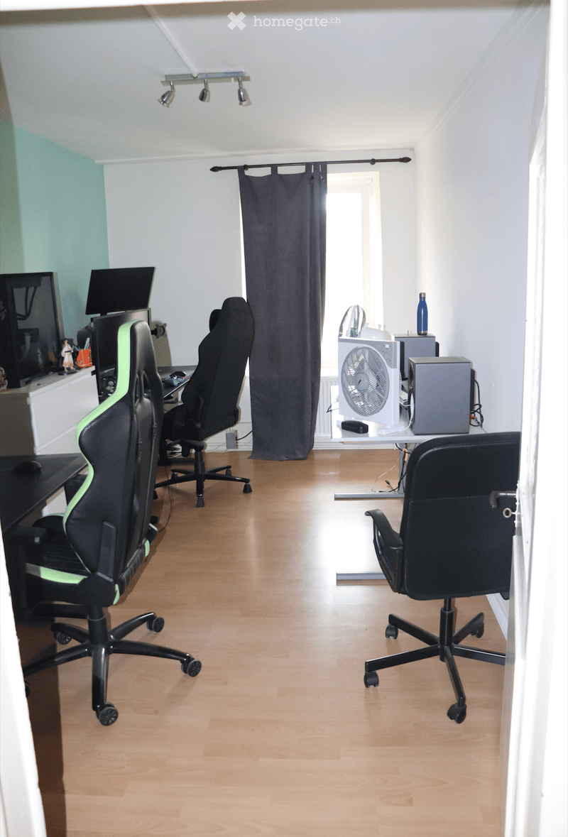 A well-lit room with wooden flooring, three black office chairs, a small table with a fan, and two screens on a desk.