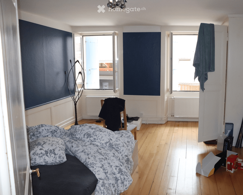 Bedroom with wooden flooring, large windows, bed with patterned bedding, chair with a jacket draped over it, coat rack, radiator, and a laptop on a small table.