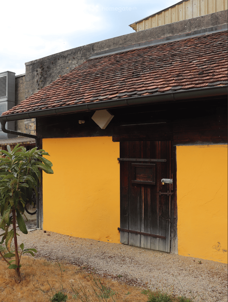 A small, single-story building with a tiled roof and a wooden door, painted in bright yellow with a gravel ground.