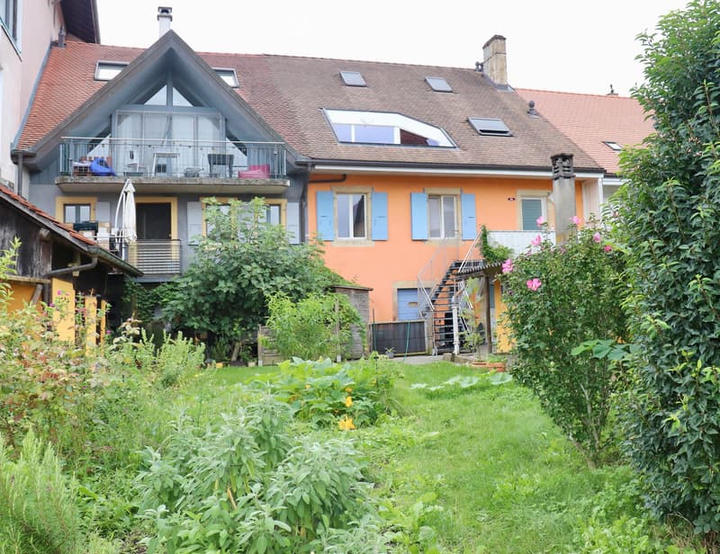 Two-story house with orange facade, multiple windows, and a balcony. The house has a well-maintained garden with various plants and a staircase leading to the entrance.