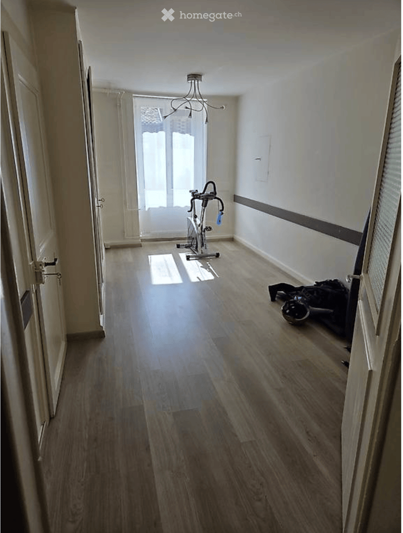 A narrow hallway with wooden flooring, a chandelier, and an exercise bike placed against the wall. The hallway has white walls and doors on either side.