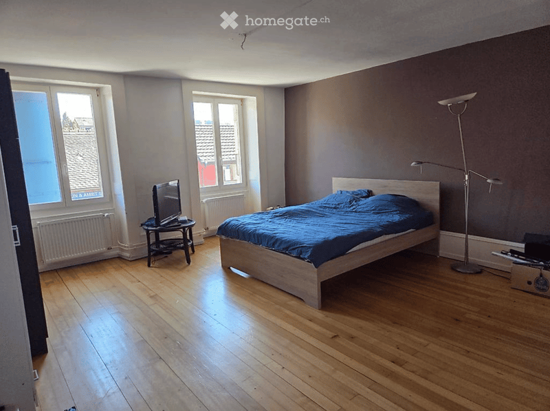 Bedroom with wooden flooring, a bed with blue bedding, a nightstand with a laptop, a small table with a TV, and a floor lamp.