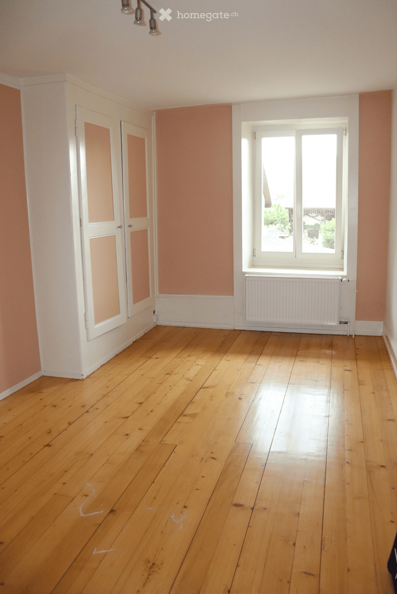 Empty room with wooden flooring, white walls, two doors, and a window with a radiator below it.