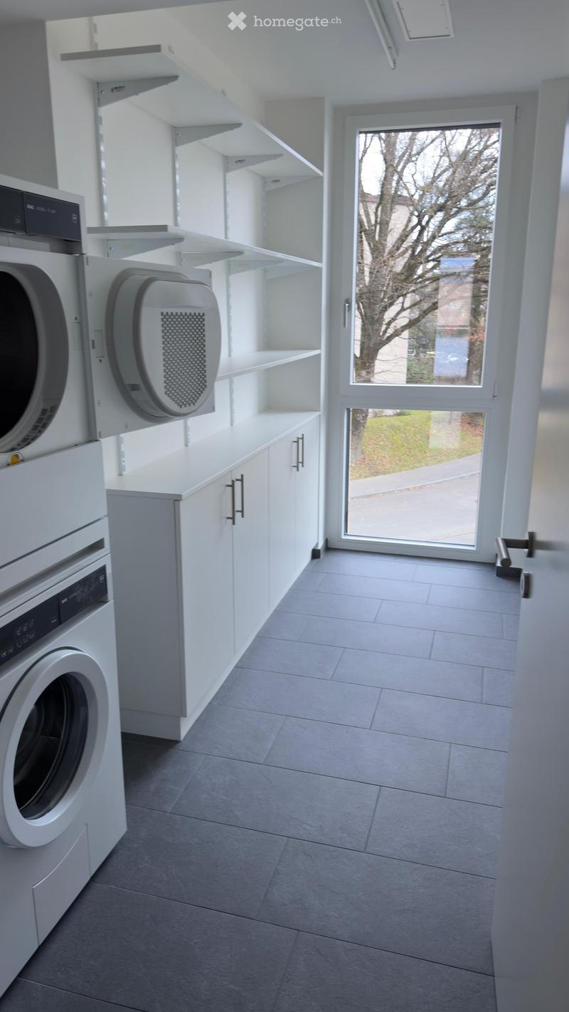 Modern bathroom with white cabinets, built-in washing machine and dishwasher, large window with view of trees.