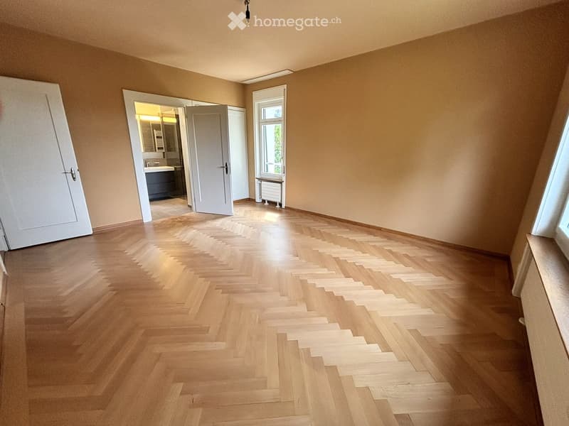 Empty room with wooden herringbone flooring, beige walls, and a large window. A door leads to a bathroom with a sink and mirror.