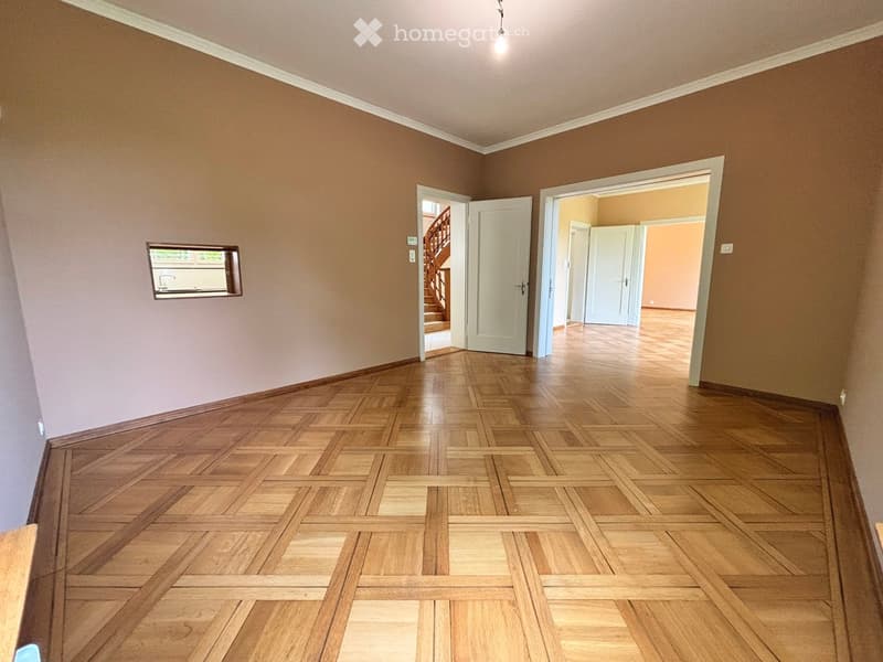 Empty hallway with wooden parquet flooring, beige walls, and white doors leading to other rooms. A small mirror is mounted on the left wall, and a staircase is visible through an open doorway.