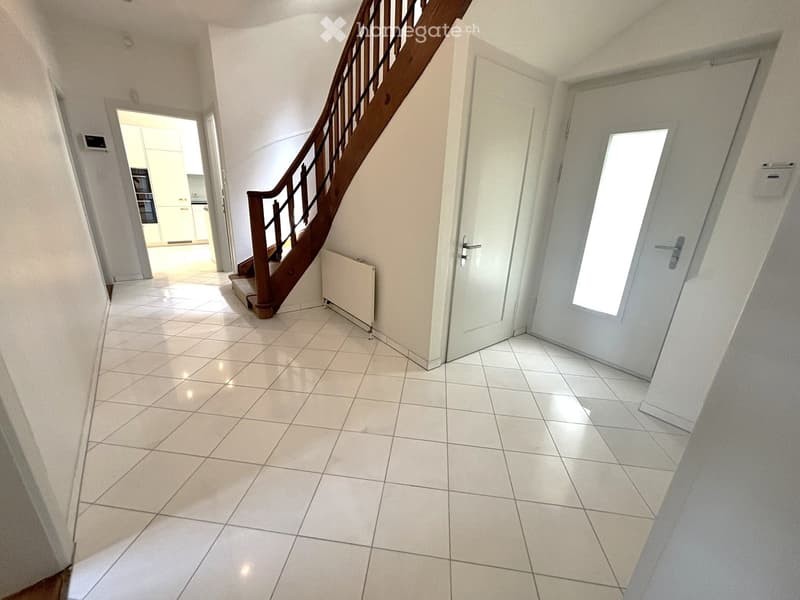 Bright hallway with tiled flooring, white walls, and a wooden staircase leading upwards. Features include a radiator, a closed door, and an open doorway leading to another room.