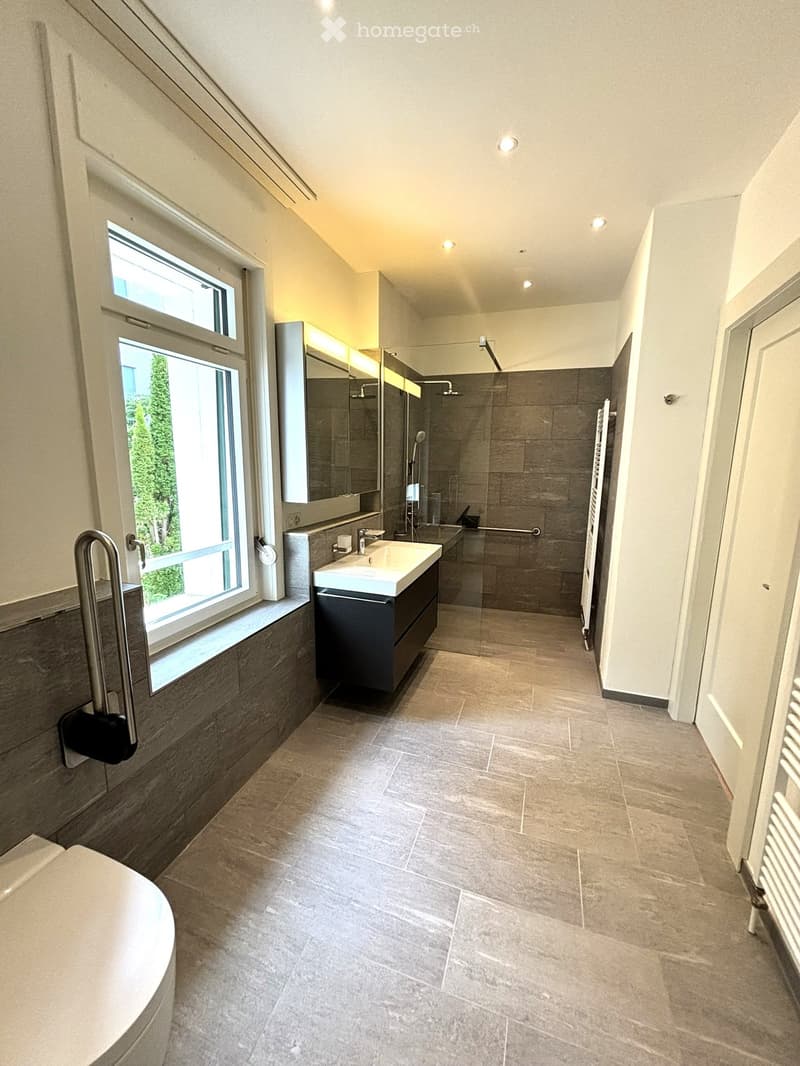 Modern bathroom with a large window, a sink with a modern faucet, a large mirror, a shower area, and a radiator. The floor is tiled with light gray tiles, and the walls are finished in a combination of light and dark tones.