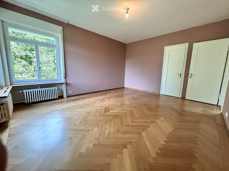 Empty room with wooden herringbone flooring, light purple walls, large window with white frame, radiator, and two white doors.